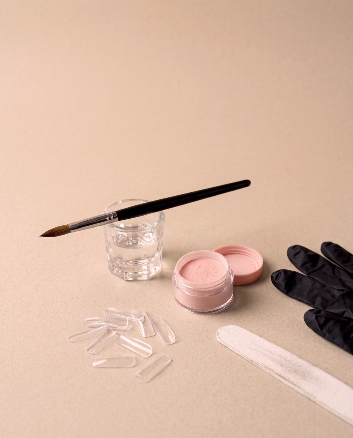 An acrylic nail bench: a brush over a dish of monomer, acrylic powder, tips, a file and black gloves